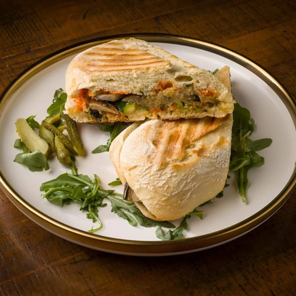 Grilled panini sandwich with vegetables, arugula, and pickles on a white plate at L'ORANGE CAFE.