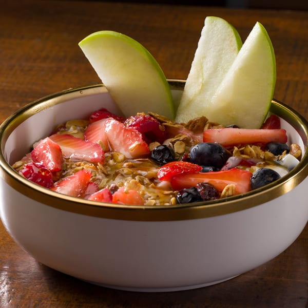 A white bowl with a gold rim filled with granola, berries, and sliced green apples.