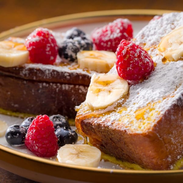 Golden French toast topped with powdered sugar, fresh raspberries, blueberries, banana slices, and syrup.